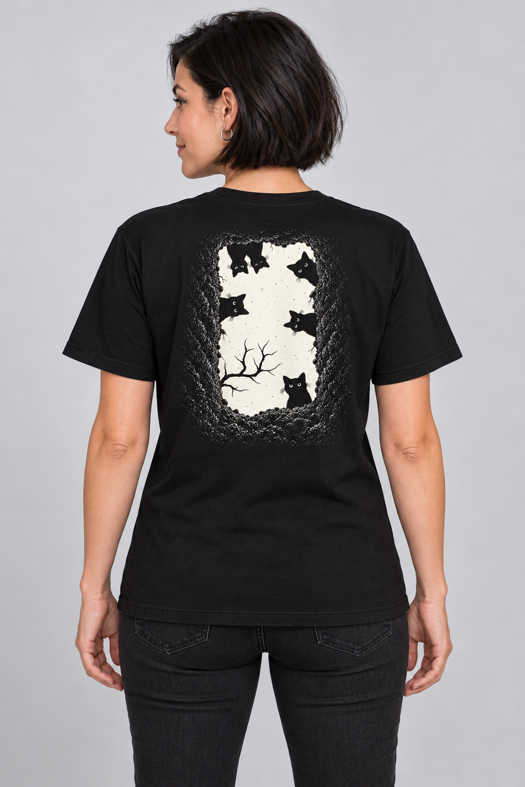 A woman shown from the back wearing a grave danger t-shirt, black graphic tee, black cat grave illustration back, gothic wordmark front design, two-sided halloween shirt, spooky cemetery graphic print, cream ink gothic lettering, torn earth night sky design, eerie cat peeking art, unisex crew neck shirt