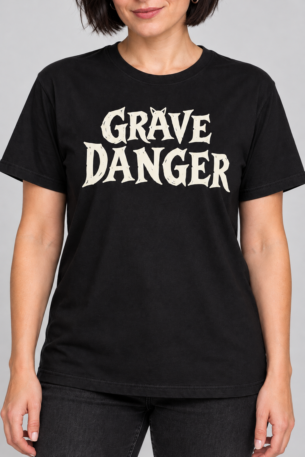 Front view of woman wearing grave danger t-shirt, black graphic tee, gothic wordmark front design, black cat grave illustration back, two-sided halloween shirt, spooky cemetery graphic print, cream ink gothic lettering, torn earth night sky design, eerie cat peeking art, unisex crew neck shirt