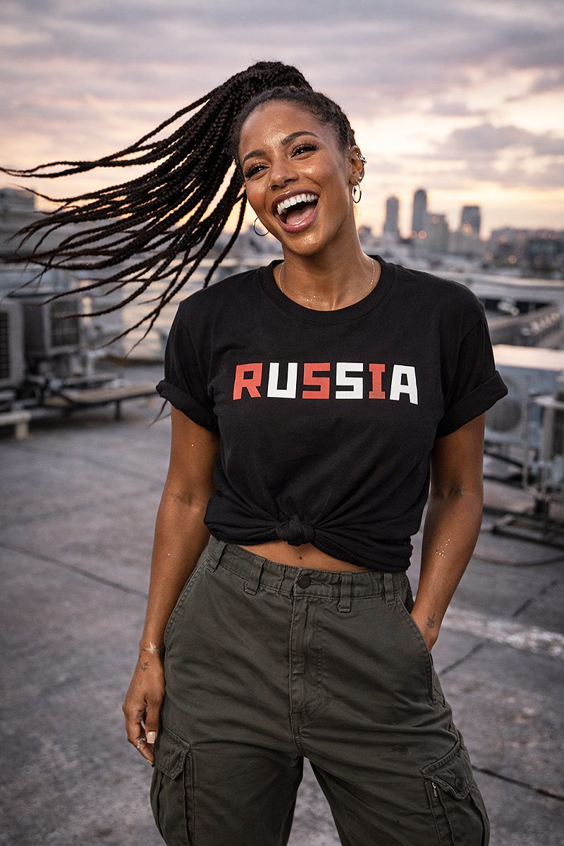 Russia USA hidden message political satire pixel font graphic tee worn by Black female model with braids windswept on rooftop at sunset black t-shirt